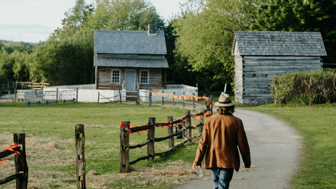 A man with a hat on walking along the path towards one of the exhibition houses at Ulster American Folk Park