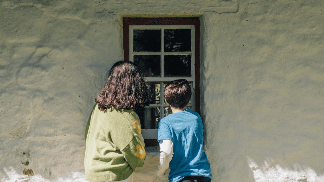 A girl in a green jumper with yellow design on the sleeve and a boy in a blue t-shirt with white long-sleeve top, both with dark hair and their backs to the camera, looking through the window of a cottage exhibit building at the Ulster American Folk Park.