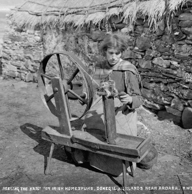 Black and white photograph of a young woman using a spinning wheel outside in rural Ireland